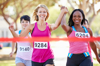 Two women running, crossing the finish line holding hands with their arms raised. Other runners in the background.