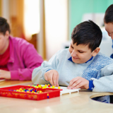 Image of 3 disabled children playing and smiling. A girl and a boy are sat at a table, playing with coloured pegboard, another boy is stood near the boy sat down, looking over his shoulder. 