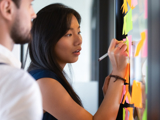 Photo focused on a woman looking at a glass board, writing notes on a post-it. There is a man standing next to her, slightly blurred.