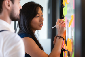 Photo focused on a woman looking at a glass board, writing notes on a post-it. There is a man standing next to her, slightly blurred.