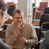 Photo of 3 people signing in a lecture hall. The man in the centre is signing, two women are communicating with him, one blurred out of focus with back to viewer.