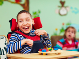 Photo of two disabled children, focused in on a boy, smiling, who is in a wheelchair, and playing with coloured bricks.