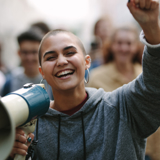 Photo focused on a smiling young woman with a megaphone in one hand and holding her other arm up. The background is blurred and of other people. 