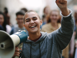 Photo focused on a smiling young woman with a megaphone in one hand and holding her other arm up. The background is blurred and of other people.