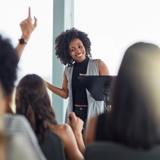 Photo of a group, with raised hands, focused in on a woman at the centre of the image happily answering questions.
