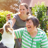 Photo focused on a middle-aged disabled woman outdoors playing with a dog, with a supporter in the background.