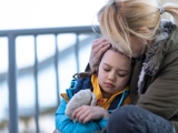 Photo of a mother, sat down in front of railings, with her daughter next to her, resting her head on her mother, who holds her head in her hand. 