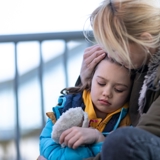 Photo of a mother, sat down in front of railings, with her daughter next to her, resting her head on her mother, who holds her head in her hand. 